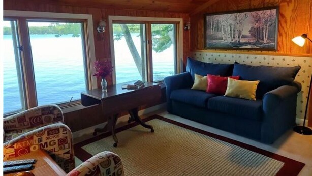 A cozy room with a blue sofa, a wooden ceiling, and a large window overlooking a body of water. - Gramp's Camp (Belgrade)