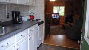 A compact kitchen with white cabinets, a coffee maker, and a red kettle. A dining area with a television is visible through an open doorway. - Hidden Cove Camp (Oakland)