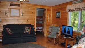 A cozy log cabin interior with a dark-colored sofa, a rocking chair, a television, and a dining area. - Hidden Cove Camp (Oakland)