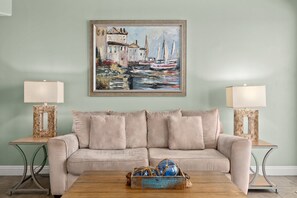 A living room with a beige sofa, two side tables with lamps, a painting of a coastal scene, and a coffee table with decorative items.