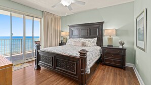 A bedroom with a large bed, a ceiling fan, a nightstand with a lamp, a dresser, and a view of the ocean through large windows. - Gulf-front condo w/ pool, hot tub and beach access (Panama City Beach)