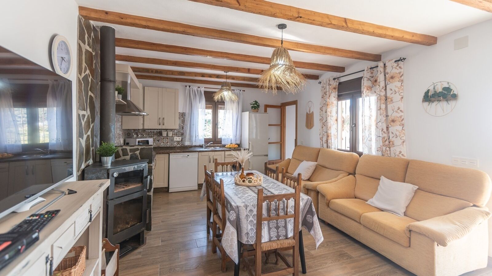 A cozy living room with a sofa and a dining table set for four. A kitchen with modern appliances and a large window is visible in the background.