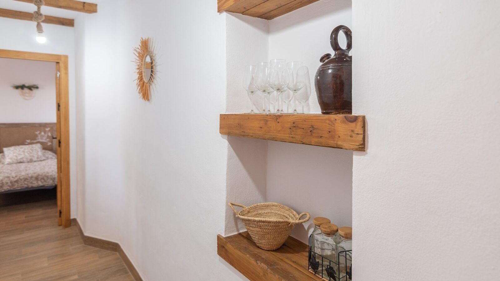 A hallway with a wooden shelf holding a woven basket, a glass vase, and a bottle. A mirror hangs on the wall, and a bed is visible in the background.