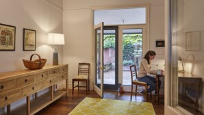 Ein Zimmer mit einem hölzernen Schminktisch, einer Lampe, einem Sessel, einem Tisch und Blick durch eine Glastür in einen Außenbereich.