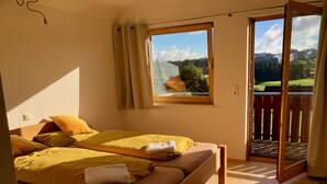 A bedroom with two beds, a window with a view of a forest and mountains, and a sliding glass door to a balcony.