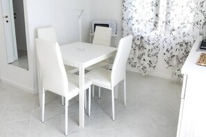 A white dining table with four chairs, a white cabinet with a microwave, and a floral curtain.