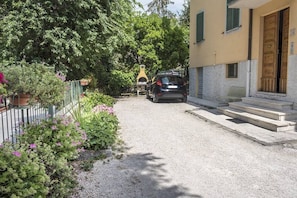 A residential street with a car parked on the side, a yellow building in the background, and a garden with pink flowers. - C004 - Camerano, new two-room apartment with air conditioning (Camerano)