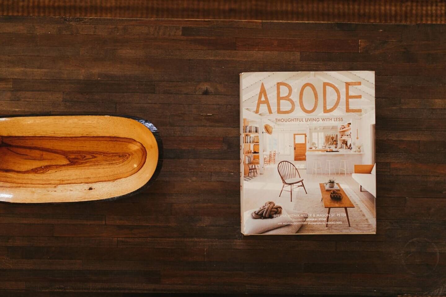 A book titled 'ABODE' with a subtitle 'THOUGHTFUL LIVING WITH LESS' on a wooden surface.