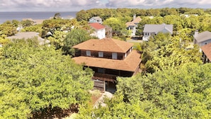 A two-story house with a red roof, surrounded by dense green trees, and a view of the sea in the background.