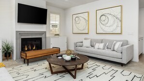 A modern living room with a fireplace, a grey sofa, a coffee table, and two abstract paintings. - GOODE.HOMES | Luxury Modern Home w/ pool/spa (Santa Clarita)