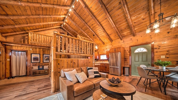 A cozy cabin interior with a wooden ceiling, a sofa, a dining table, and a staircase leading to an upper level. - Cambridge Springs Cabin Near French Creek! (Cambridge Springs)