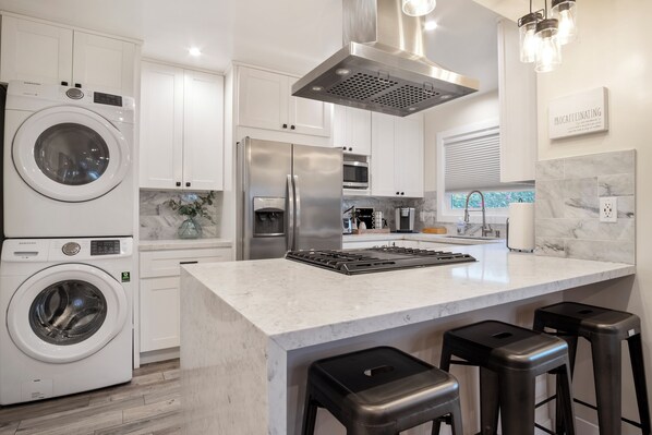 A modern kitchen with a built-in washer, stainless steel appliances, a marble countertop, and bar stools.