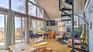 Een ruime woonkamer met een groot raam, een houten vloer, een bureau, een stoel, een bank en een spiltrap.