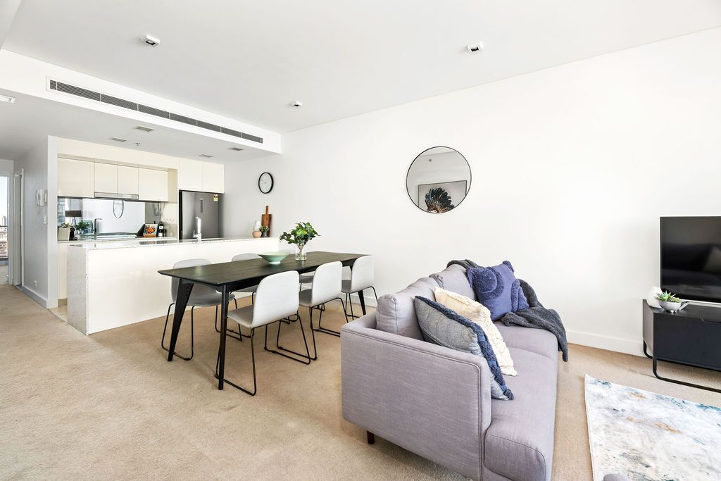 A modern living room with a grey sofa, a dining table with chairs, a kitchen area, and a TV stand.
