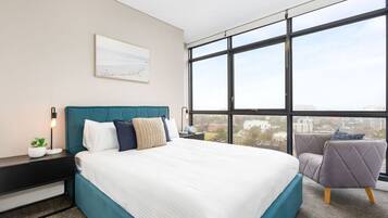 A modern bedroom with a large bed, a view of the city through large windows, and a small seating area.