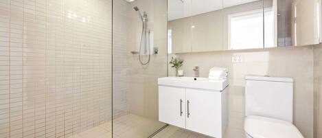 Un baño moderno con ducha de vidrio, azulejos blancos, un lavamanos blanco y un inodoro.