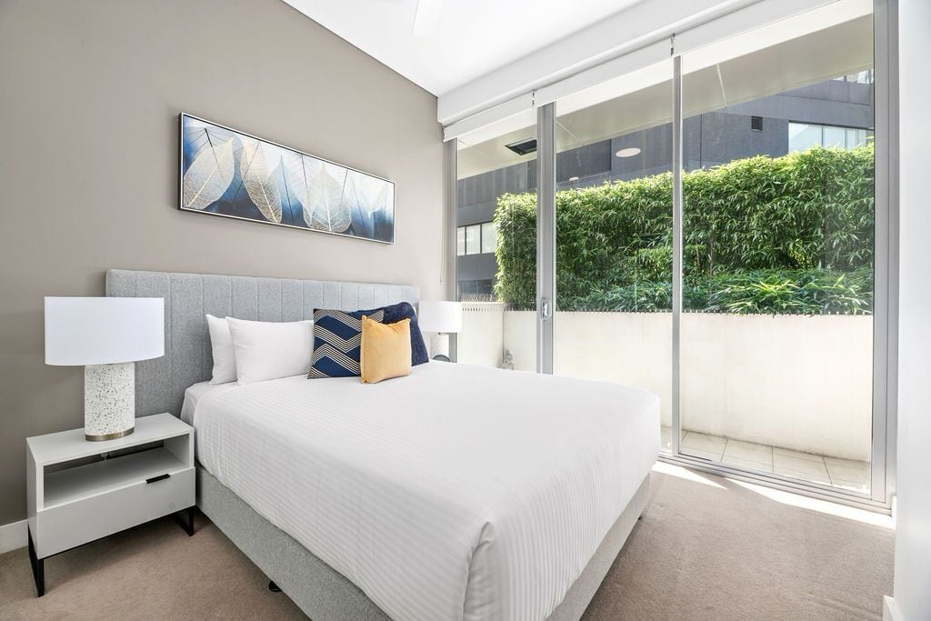 A modern bedroom with a large bed, a nightstand, a wall-mounted artwork, and a view of a green hedge through a sliding glass door.