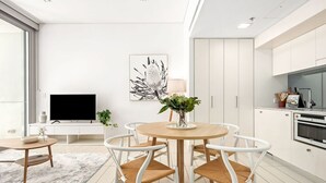 A modern living room with a round wooden table, white chairs, a flat-screen TV, a painting, and a kitchen area with cabinets and appliances. - The Alta Apartments by Urban Rest (Surry Hills)