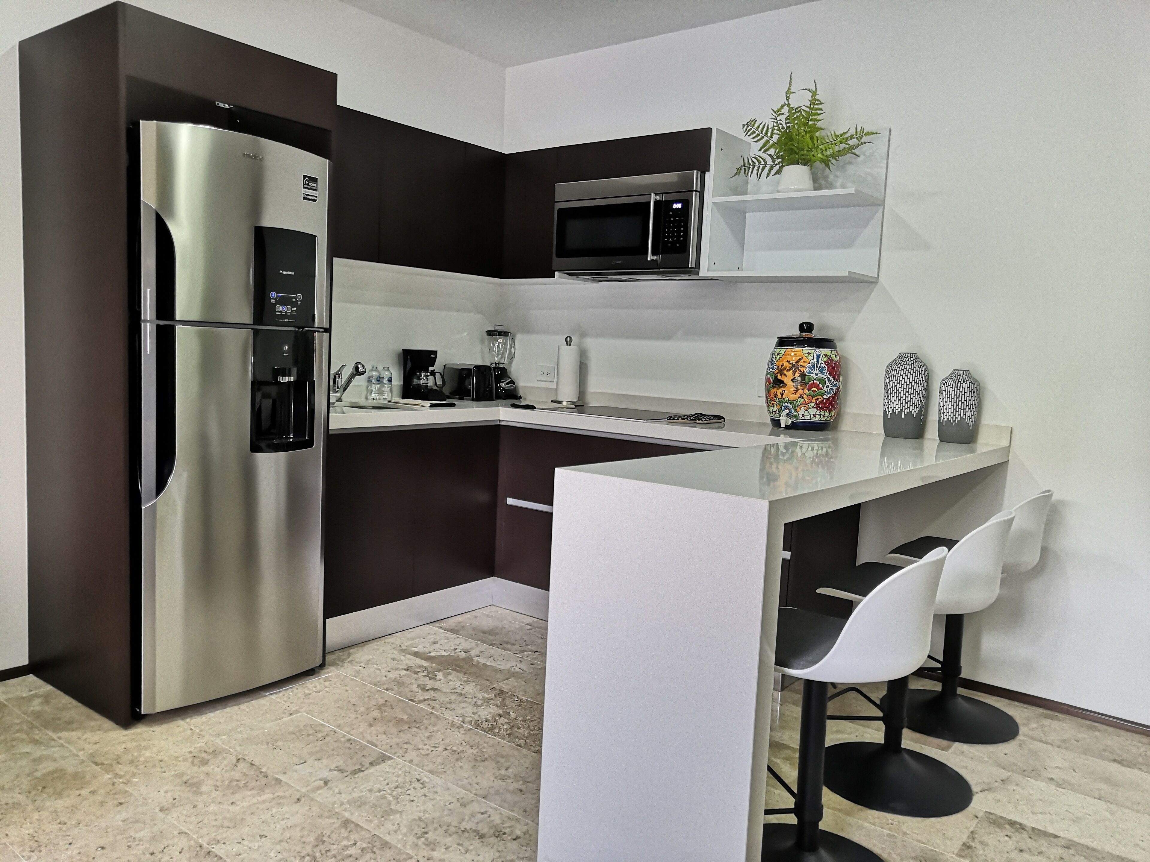 A modern kitchen with a stainless steel refrigerator, a microwave, and a coffee maker. There is a small bar area with two white chairs.