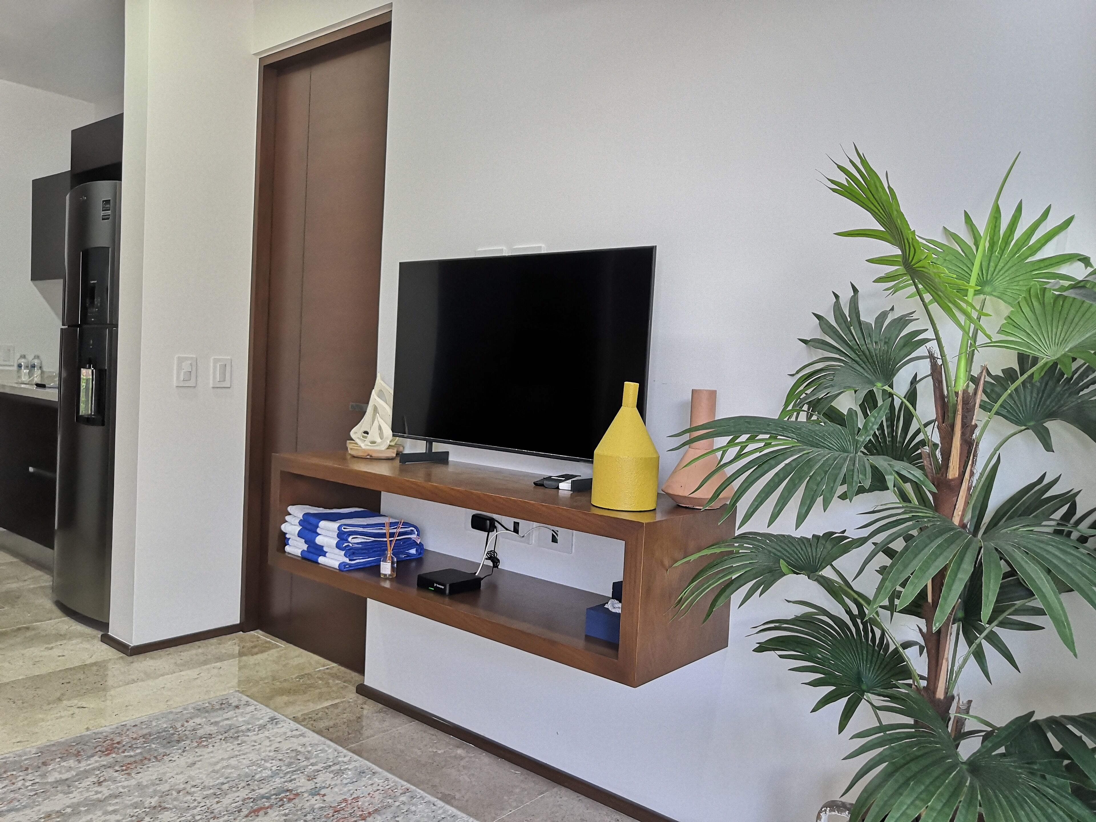 Un salon moderne avec un téléviseur à écran plat posé sur un meuble en bois, une petite plante et une serviette pliée sur le sol.