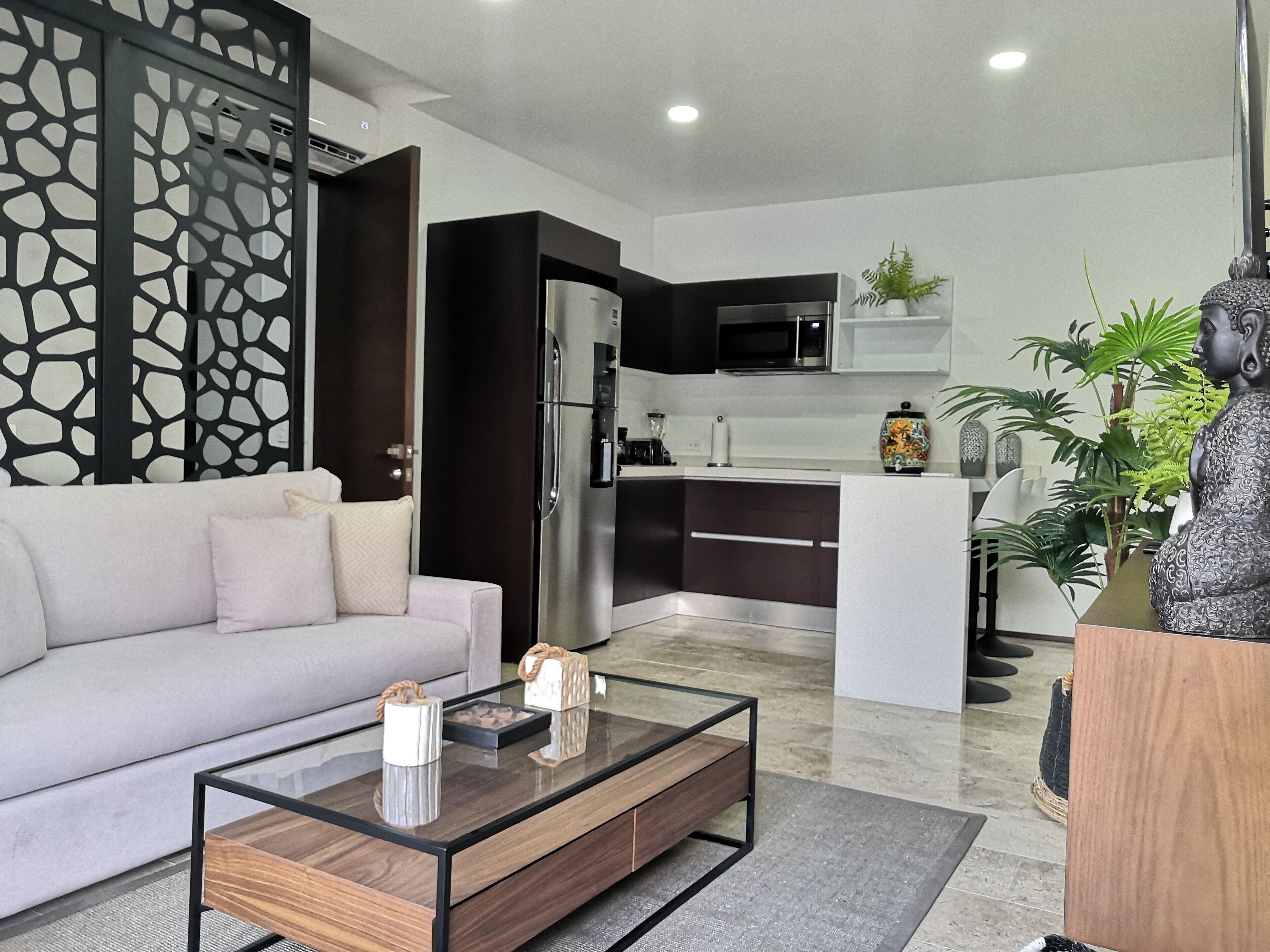 Ein modernes Wohnzimmer mit einer Couch, einem Couchtisch und Dekoration. Im Hintergrund ist eine Küche mit Kühlschrank und Mikrowelle zu sehen.