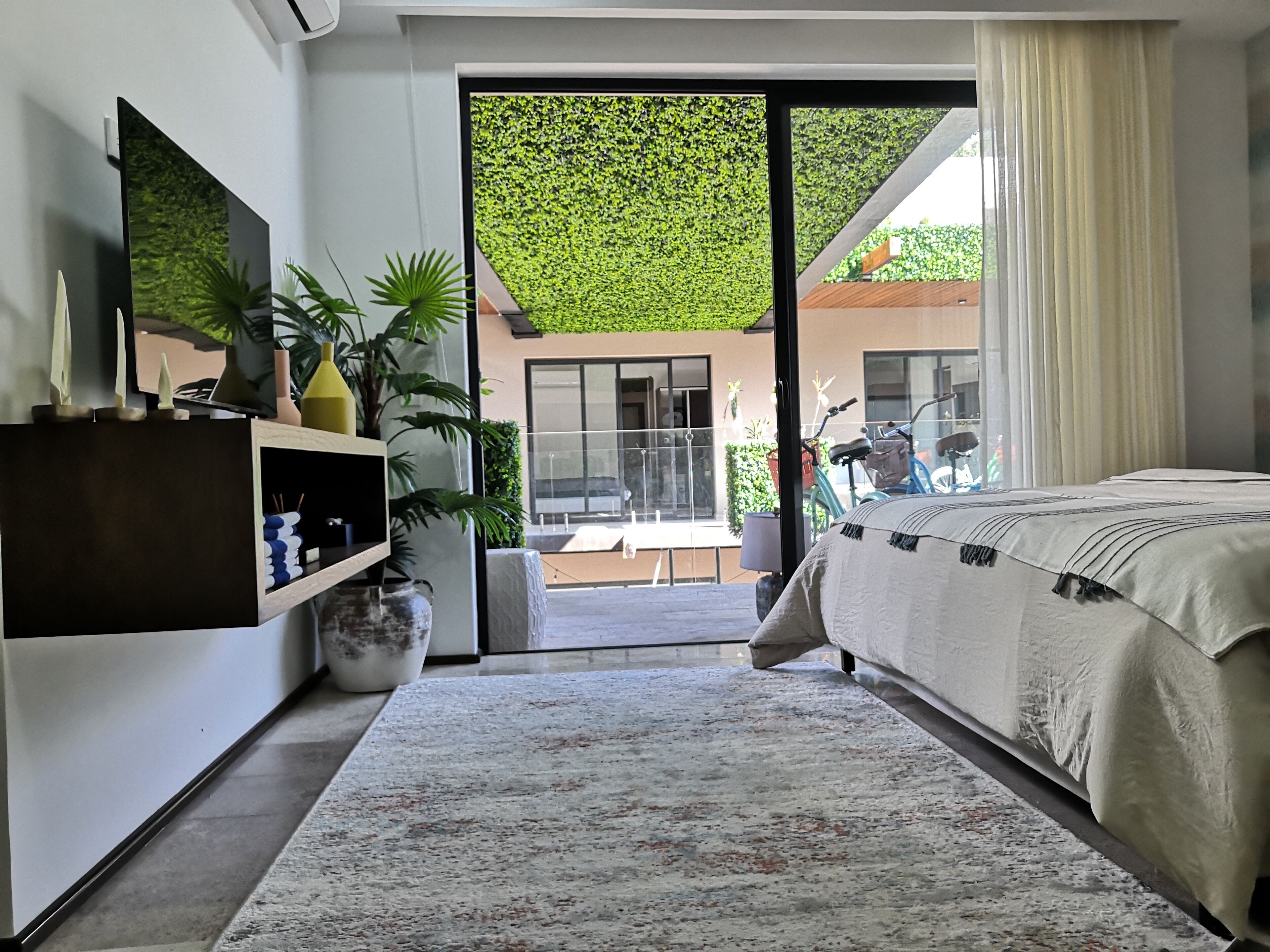 Un dormitorio moderno con una cama grande, un televisor montado en la pared, una puerta corrediza de vidrio que da a un balcón y un pequeño estante con objetos decorativos.