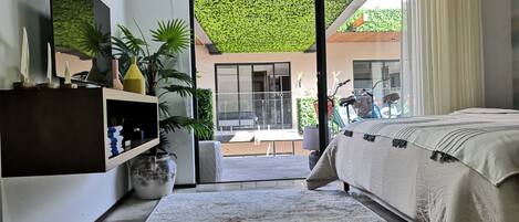 Un dormitorio moderno con una cama grande, un televisor montado en la pared, una puerta corrediza de vidrio que da a un balcón y un pequeño estante con objetos decorativos.