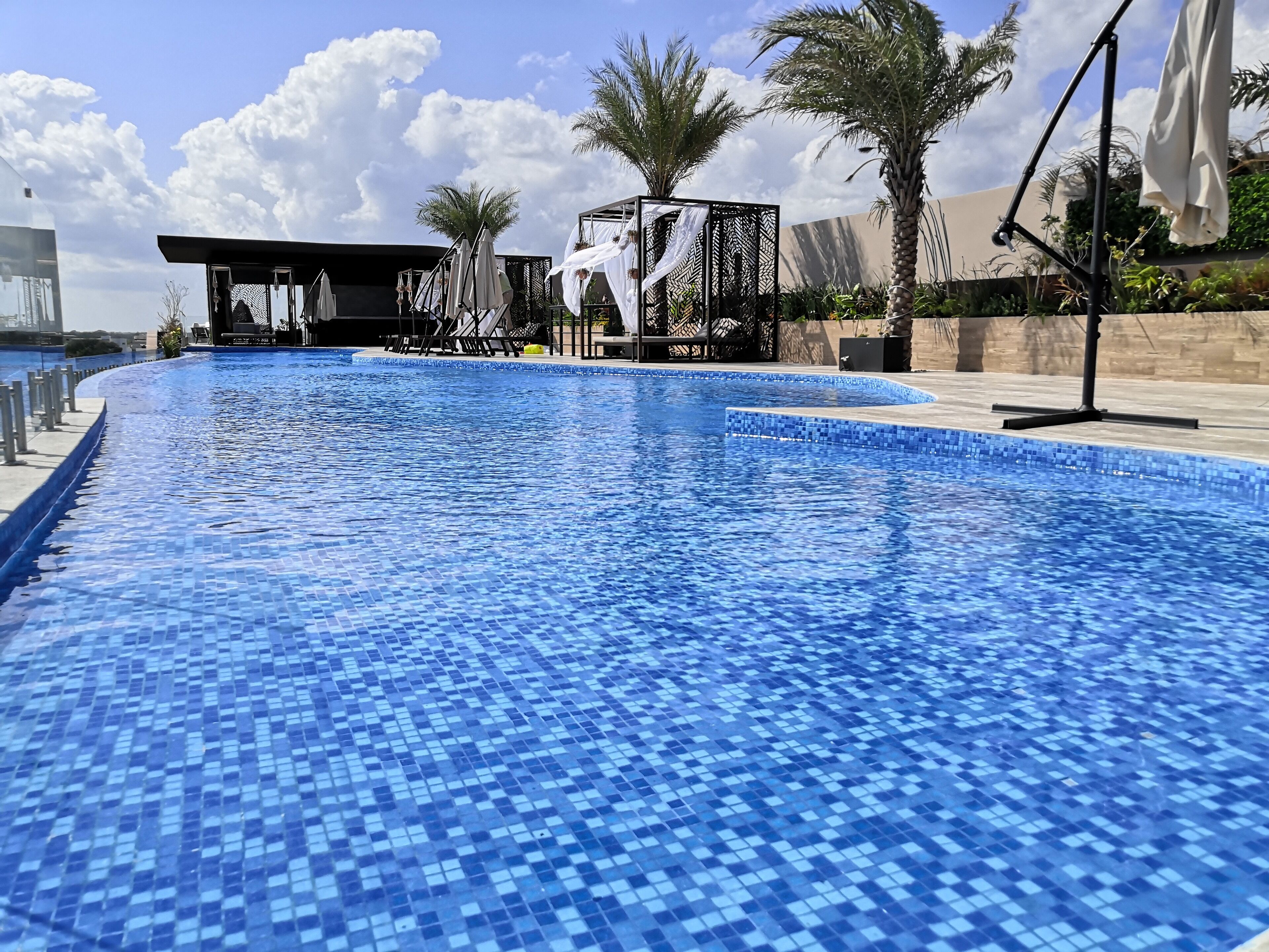 A swimming pool with blue mosaic tiles, surrounded by palm trees and a covered seating area.