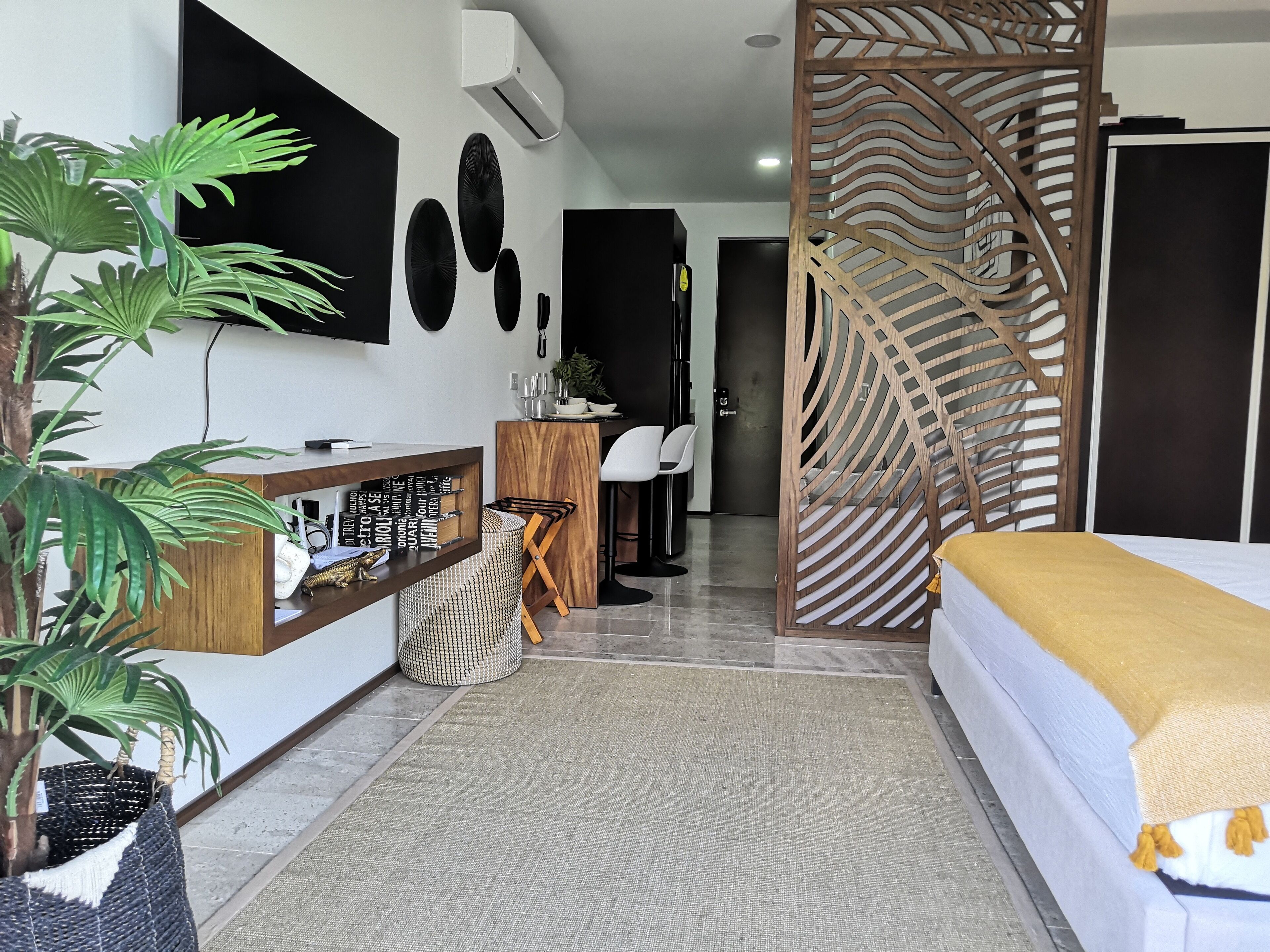 A modern hotel room with a bed, a desk, a chair, a decorative wooden partition, and a potted plant.