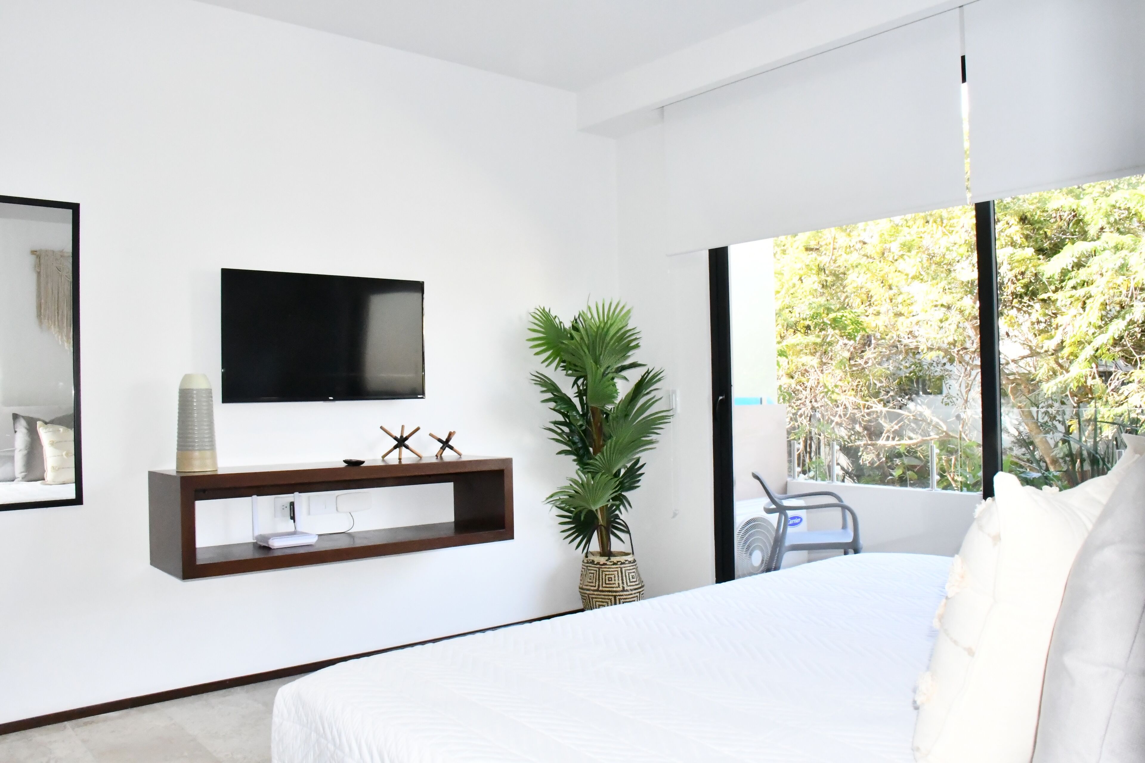 A modern bedroom with a large bed, a TV mounted on the wall, a potted plant, and a window with a view of trees.