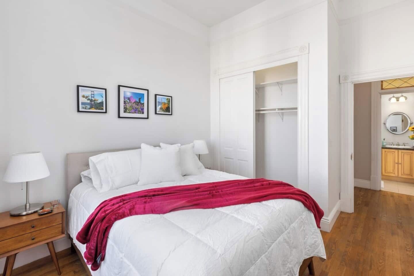 A bedroom with a bed, a nightstand, a lamp, a wardrobe, and a bathroom visible through an open door.
