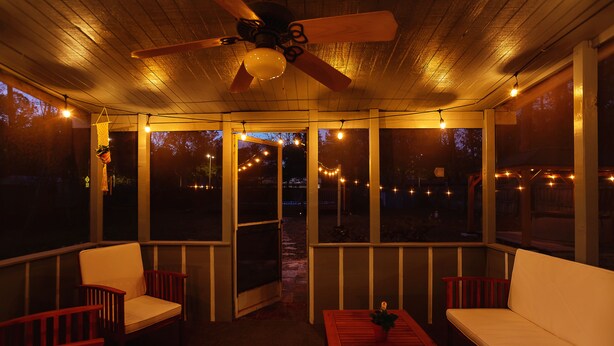 Une terrasse couverte, aménagée avec des meubles en bois, des guirlandes lumineuses et un ventilateur de plafond.