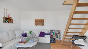 Een woonkamer met een witte bank, een salontafel, een klein houten rekje, een kleurrijk abstract schilderij en een houten ladder.