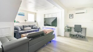 A modern living room with a grey sofa, a flat-screen TV, a desk with a chair, and a window with blinds.