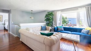 A living room with a beige sofa, a blue sofa, and a small table. There is a plant and a window with curtains.