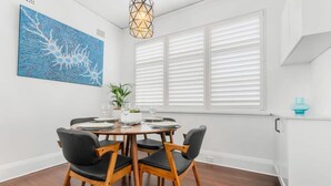 A dining area with a round table, black chairs, white walls, a blue artwork, and a window with blinds. - Coogee Riviera in Coogee (Coogee)