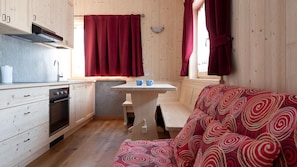 A compact kitchen with wooden cabinets, a built-in oven, and a sink. There is a small table with two blue cups and a red patterned sofa.