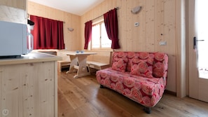 A cozy cabin interior with a wooden floor, a sofa with a red and white pattern, a wooden dining table, and a window with red curtains.