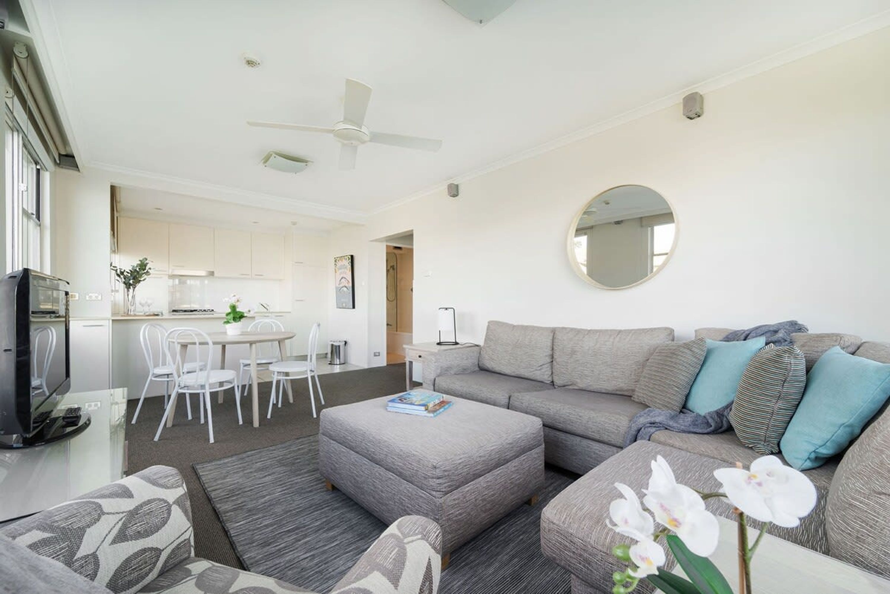 A modern living room with a grey sofa, ottoman, and a dining area with white chairs and a table.