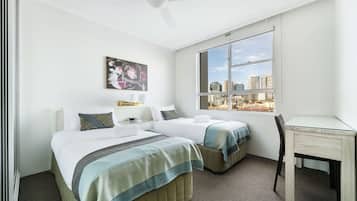Habitación de hotel con dos camas, un escritorio, una silla, un ventilador de techo, una ventana con vista a la ciudad y un cuadro en la pared.