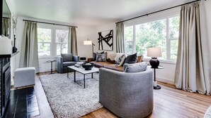A modern living room with a sofa, armchair, and coffee table. There is a fireplace, a lamp, and a large window with curtains. - Stunning U of O Home – Just Steps from Hayward Field! (Eugene)