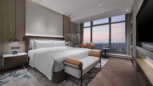 Una habitación de hotel moderna con una cama grande, zona de estar, televisión y vistas a la ciudad a través de amplias ventanas.