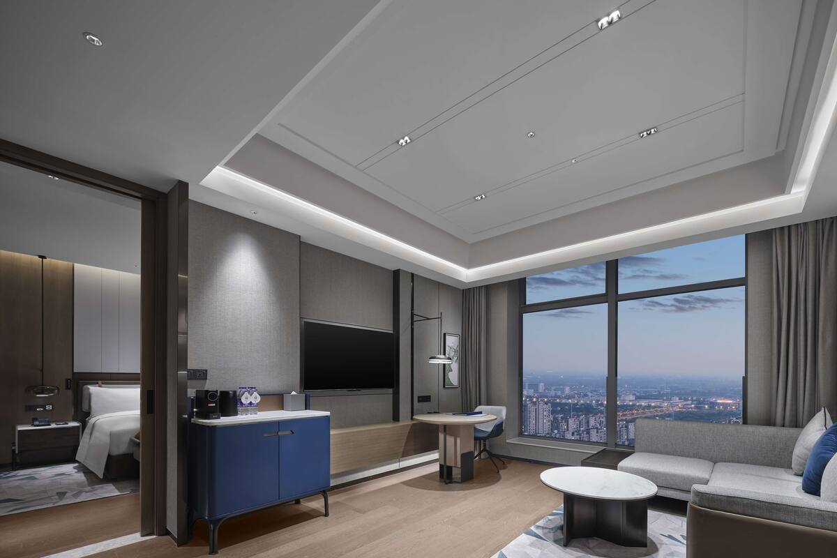 A modern hotel room with a large window offering a city view, a flat-screen TV, a blue minibar, a round coffee table, and a sofa.