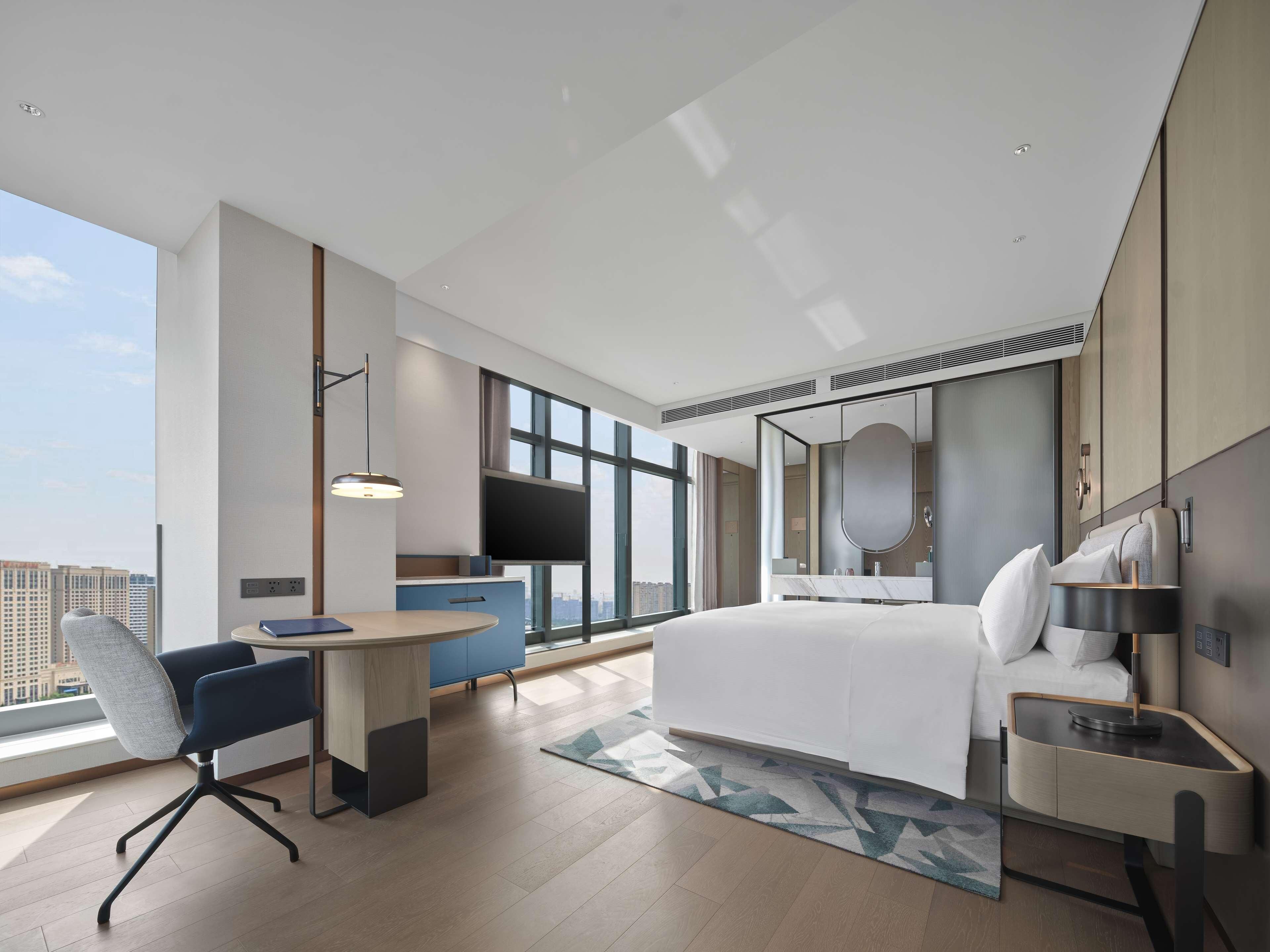 Een moderne hotelkamer met een groot bed, een bureau, een stoel, een kleine tafel, een groot raam en uitzicht op de stad.