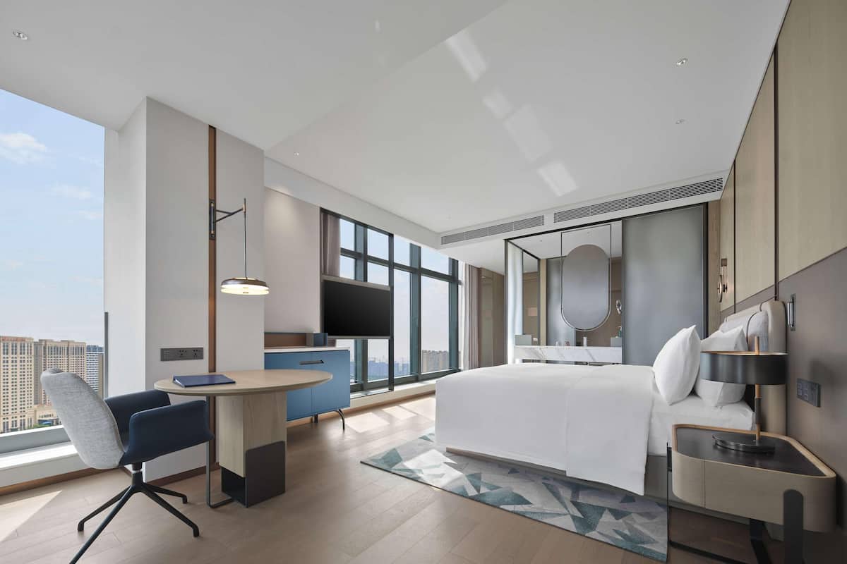 A modern hotel room with a large bed, a desk, a chair, a small table, a large window, and a city view.