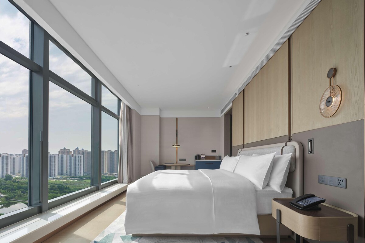 Una habitación de hotel moderna con un ventanal grande, una cama, una mesita de noche con un teléfono y un escritorio con una lámpara.
