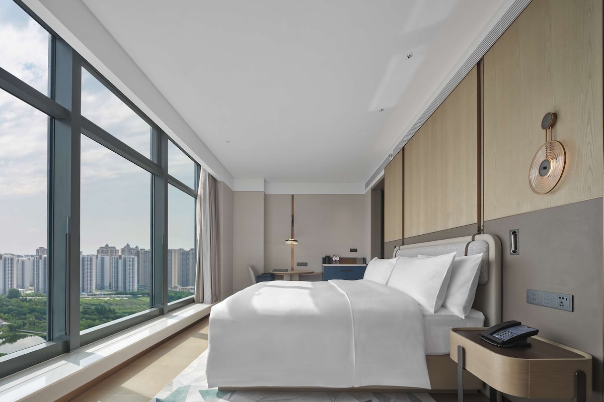 A modern hotel room with a large window, a bed, a bedside table with a phone, and a desk with a chair.
