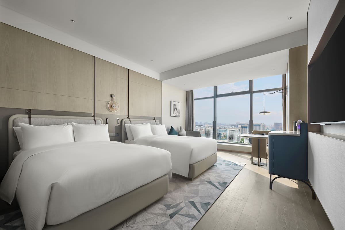 A modern hotel room with two beds, a desk, and a large window offering a city view.
