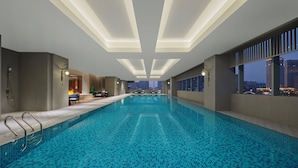 Piscina interior moderna con azulejos azul claro, iluminación empotrada y vistas a la ciudad a través de amplias ventanas.