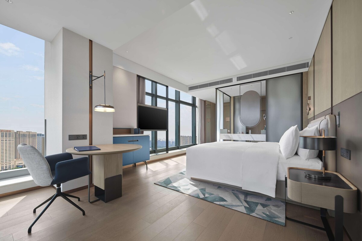 Una habitación de hotel moderna con una cama grande, un escritorio, una silla, una mesita pequeña, un ventanal amplio y vistas a la ciudad.
